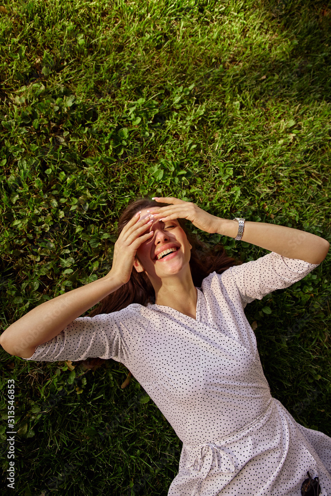 Happy young attractive brunette female in elegant dress raising hands to her face while laughing joyfully with closed eyes, lying outdoor on warm spring day Happy young attractive brunette female in elegant dress raising hands to her face while laughing joyfully with closed eyes, lying outdoor on warm spring day