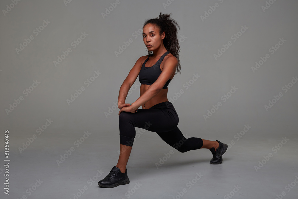 Attractive young curly brunette female with dark skin looking at camera with concentrated face while stretching her legs over grey background. Fitness model exercising indoors Attractive young curly brunette female with dark skin looking at camera with concentrated face while stretching her legs over grey background. Fitness model exercising indoors