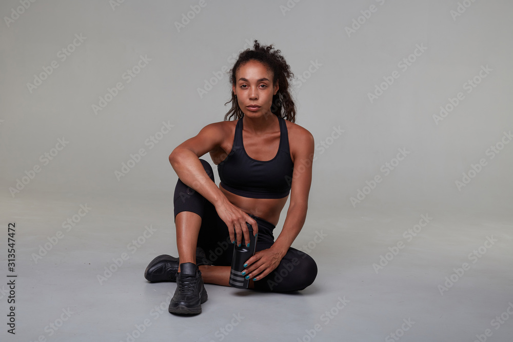 Serious young sporty dark skinned curly brunette woman with casual hairstyle looking at camera with folded lips while sitting on floor with crossed legs, isolated over grey background Serious young sporty dark skinned curly brunette woman with casual hairstyle looking at camera with folded lips while sitting on floor with crossed legs, isolated over grey background