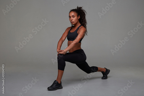 Attractive young curly brunette female with dark skin looking at camera with concentrated face while stretching her legs over grey background. Fitness model exercising indoors