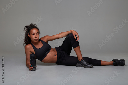 Athletic young attractive curly brunette woman with dark skin drinking water from fitness bottle while training indoor, posing against grey background in sporty clothes