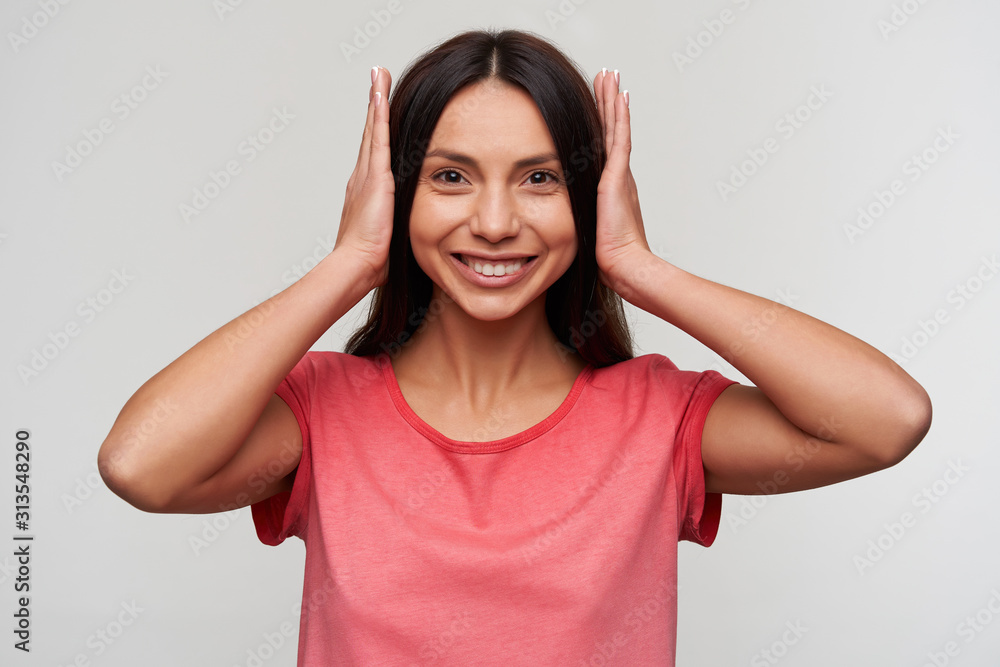 Cheerful young lovely dark haired lady with natural makeup closing her ears with hands and looking happily at camera and smiling widely, standing against white background Cheerful young lovely dark haired lady with natural makeup closing her ears with hands and looking happily at camera and smiling widely, standing against white background
