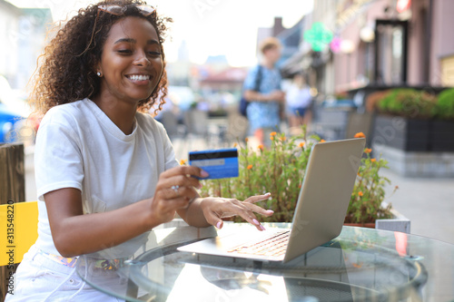 Happy attractive woman using credit card and laptop in cafe outdoors.