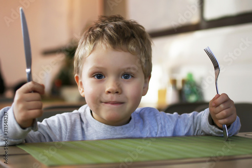 A little smiling boy with cutlery in his hands, sits at a table in a cafe and waits for his order.Concept children's diet and nutrition.