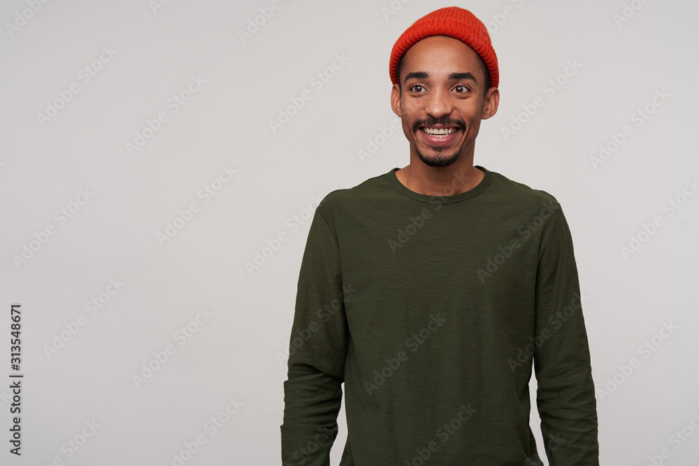 Positive young lovely bearded dark skinned brunette male in red hat and casual clothes looking aside with charming smile while standing over white background with hands down
