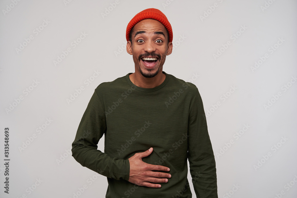 Portrait of overjoyed young bearded dark skinned guy looking at camera with wide eyes opened and holding hand on his belly while laughing happily, isolated over white background Portrait of overjoyed young bearded dark skinned guy looking at camera with wide eyes opened and holding hand on his belly while laughing happily, isolated over white background