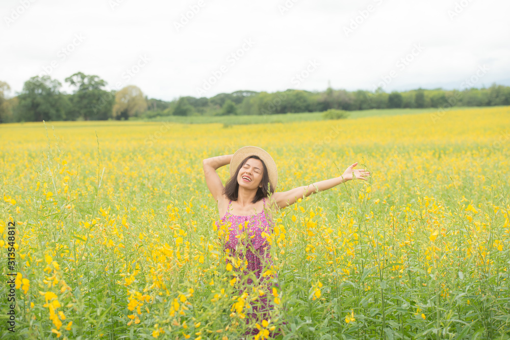 asia woman with a hat in her hand walks in a field with field flowers and smiles sincerely, happy enjoying summer in yellow field at sunset. smiling with arms raised up.  concept of freedom.