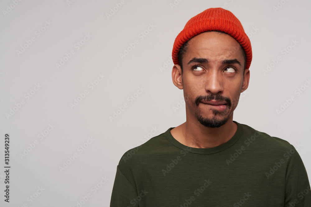 Puzzled young pretty dark skinned bearded brunette man biting underlip while looking upwards, plotting something, standing against white background in casual wear Puzzled young pretty dark skinned bearded brunette man biting underlip while looking upwards, plotting something, standing against white background in casual wear