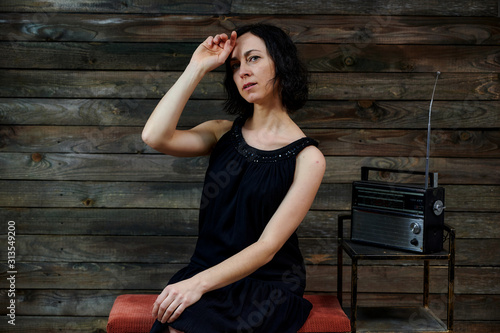 Portrait of a cute emotional brunette woman in a black dress sitting on a chair on an alternative gray background in vintage interior with a radio on a stand.