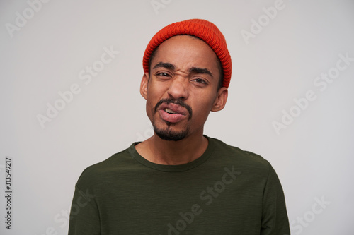 Displeased young pretty bearded dark skinned brunette guy twisting his mouth and looking at camera with pout, wearing red hat and khaki sweater while standing over white background © timtimphoto
