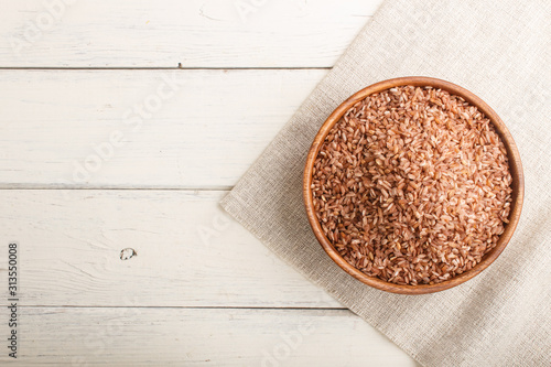 Wooden bowl with unpolished brown rice on a white wooden background. Top view, copy space.