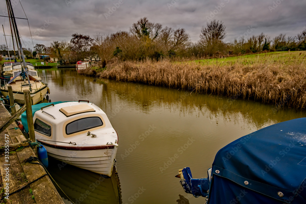 Fototapeta premium Old motor boats and cruisers moored up in a dyke off the Norfolk Broads on a dull and overcast winter's day
