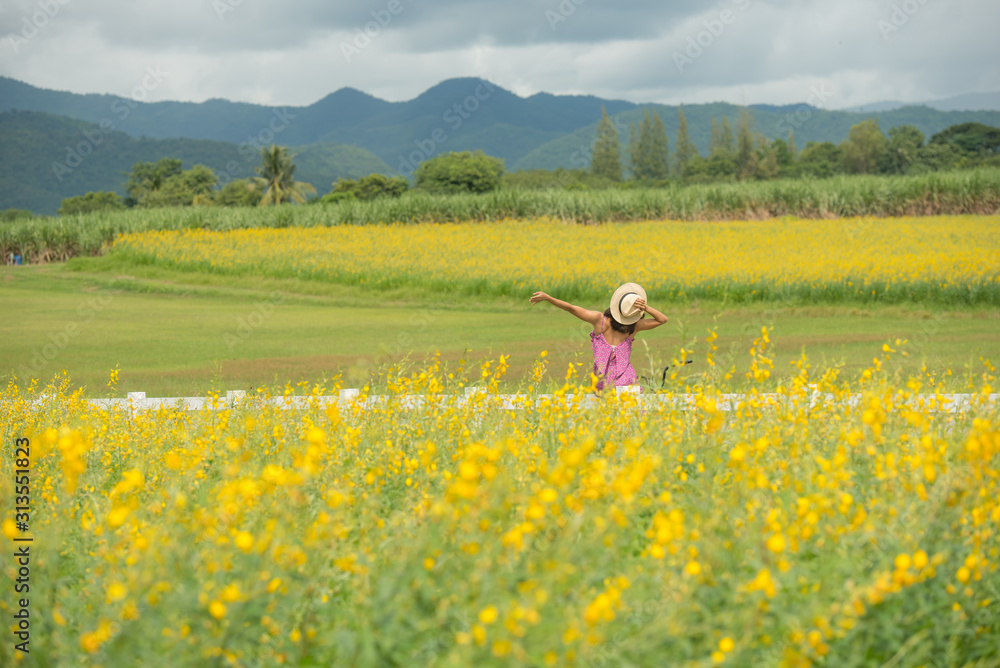 asia woman with a hat in her hand walks in a field with field flowers and smiles sincerely, happy enjoying summer in yellow field at sunset. smiling with arms raised up.  concept of freedom.