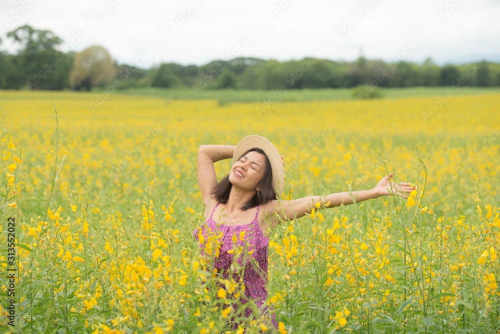 asia woman with a hat in her hand walks in a field with field flowers and smiles sincerely, happy enjoying summer in yellow field at sunset. smiling with arms raised up.  concept of freedom.