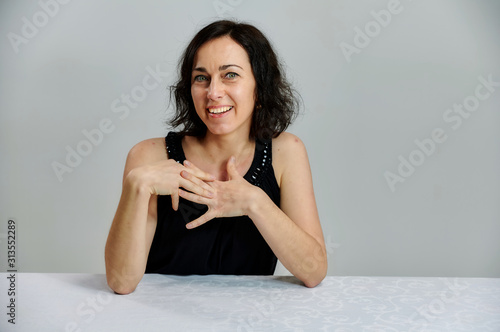 Model Sits at a table right in front of the camera with vivid emotions. Portrait of a cute smiling talking brunette woman in a black dress on a white background.