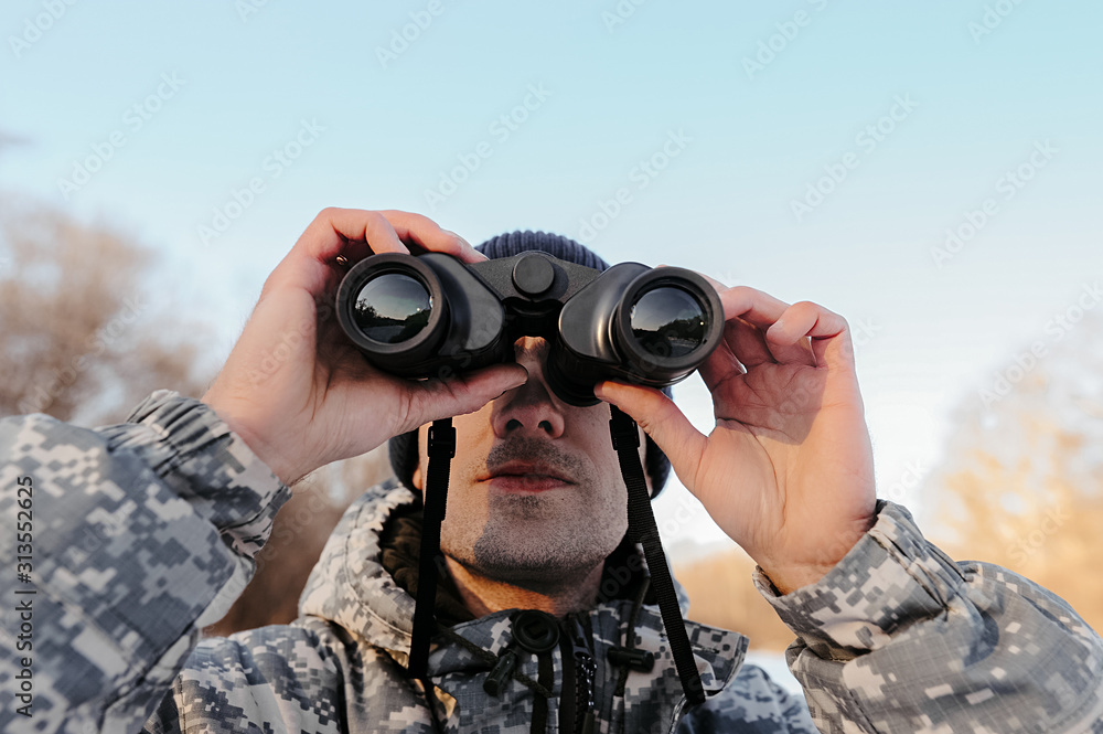 close-up face of a man with bristles and camouflage clothing, a tourist looks at winter nature through binoculars close-up face of a man with bristles and camouflage clothing, a tourist looks at winter nature through binoculars