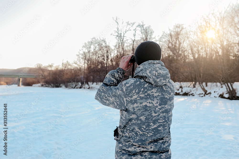 hunters back in military clothes and a blue hat looks through binoculars, the discovery of winter wildlife by man hunters back in military clothes and a blue hat looks through binoculars, the discovery of winter wildlife by man