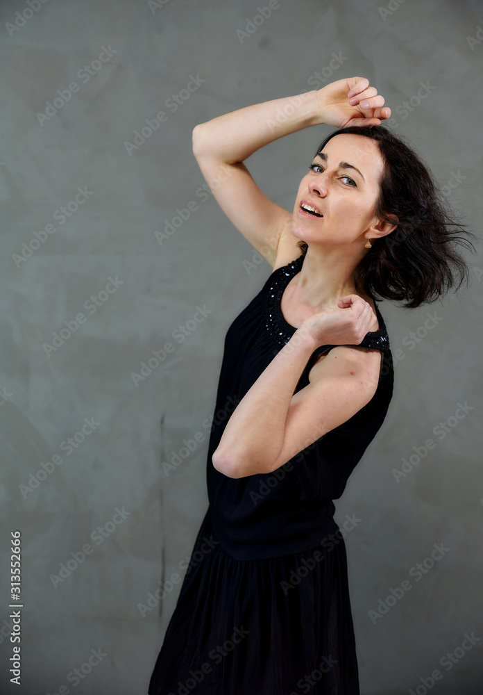 Model Standing right in front of the camera with vivid emotions. Vertical portrait of a cute smiling brunette woman in black dress on gray alternative background. Model Standing right in front of the camera with vivid emotions. Vertical portrait of a cute smiling brunette woman in black dress on gray alternative background.