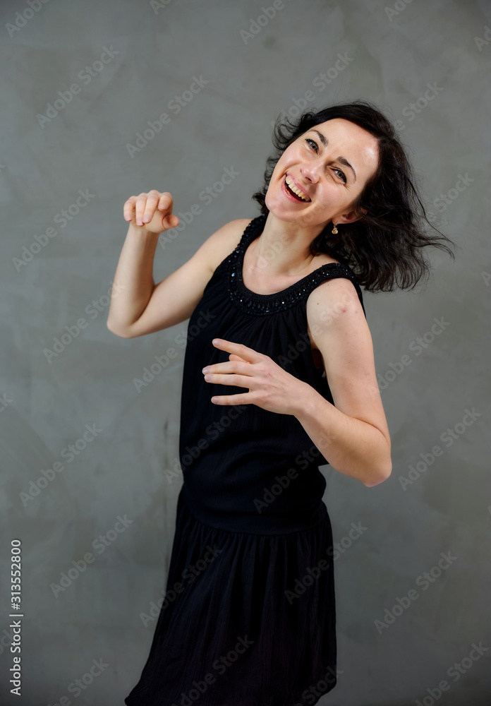 Model Standing right in front of the camera with vivid emotions. Vertical portrait of a cute smiling brunette woman in black dress on gray alternative background. Model Standing right in front of the camera with vivid emotions. Vertical portrait of a cute smiling brunette woman in black dress on gray alternative background.