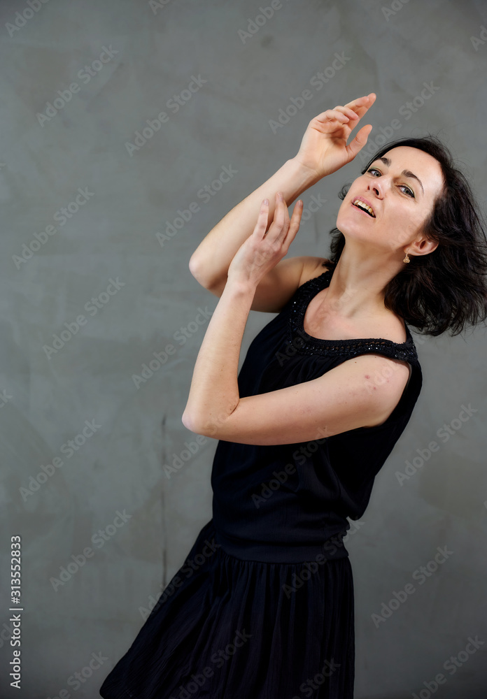 Model Standing right in front of the camera with vivid emotions. Vertical portrait of a cute smiling brunette woman in black dress on gray alternative background.
