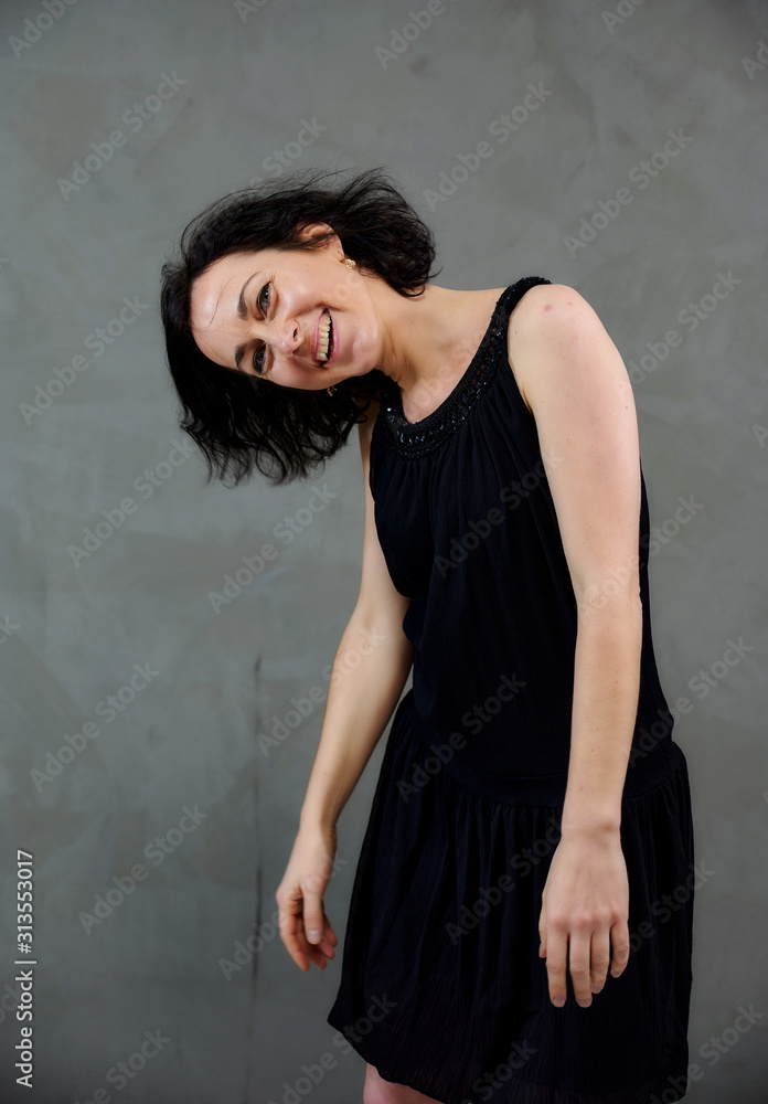 Model Standing right in front of the camera with vivid emotions. Vertical portrait of a cute smiling brunette woman in black dress on gray alternative background. Model Standing right in front of the camera with vivid emotions. Vertical portrait of a cute smiling brunette woman in black dress on gray alternative background.