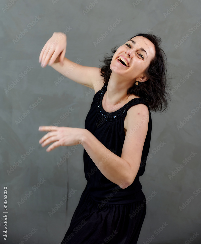 Model Standing right in front of the camera with vivid emotions. Vertical portrait of a cute smiling brunette woman in black dress on gray alternative background. Model Standing right in front of the camera with vivid emotions. Vertical portrait of a cute smiling brunette woman in black dress on gray alternative background.