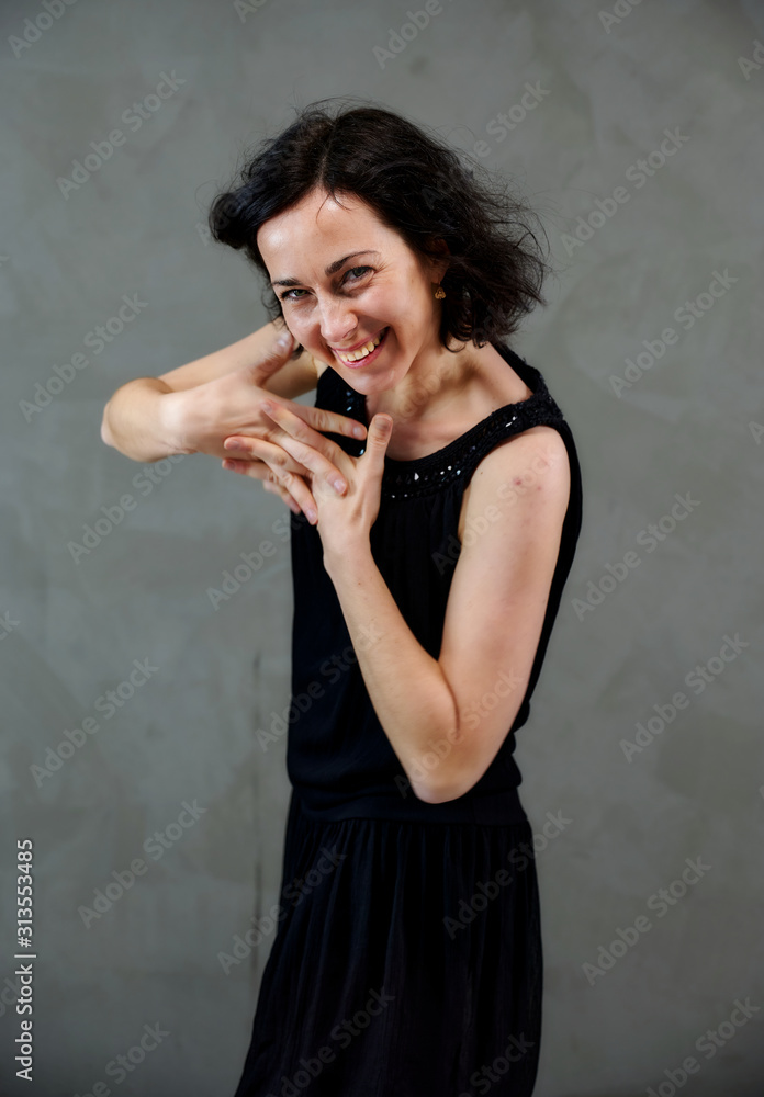 Fototapeta premium Model Standing right in front of the camera with vivid emotions. Vertical portrait of a cute smiling brunette woman in black dress on gray alternative background.