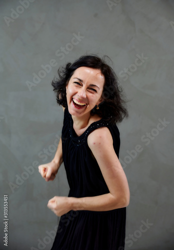 Model Standing right in front of the camera with vivid emotions. Vertical portrait of a cute smiling brunette woman in black dress on gray alternative background.