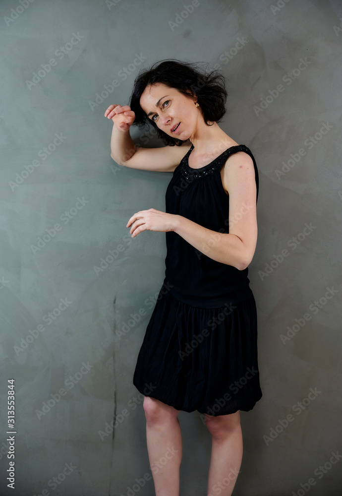 Model Standing right in front of the camera with vivid emotions. Vertical portrait of a cute smiling brunette woman in black dress on gray alternative background. Model Standing right in front of the camera with vivid emotions. Vertical portrait of a cute smiling brunette woman in black dress on gray alternative background.