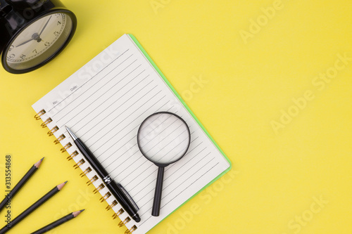 Education concept. Flat lay composition of open notebook, pen, magnifying glass, pencils and alarm clock. Copy space over green background. Top view on various stationery on desk. Creative workspace.
