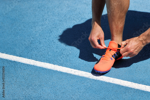 Athlete sprinter getting ready to run tying up shoe laces on stadium running tracks. Man runner preparing for cardio training outdoors. Fitness and sports. Blue background.