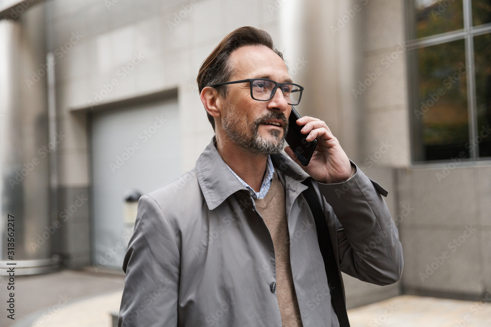 Image of pleased businessman talking on cellphone while standing Image of pleased businessman talking on cellphone while standing