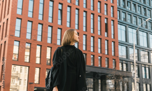 Fashionable girl in stylish casual clothes stands on the street against the backdrop of modern architecture and looks away. Street photo of fashionable lady in dark clothing.