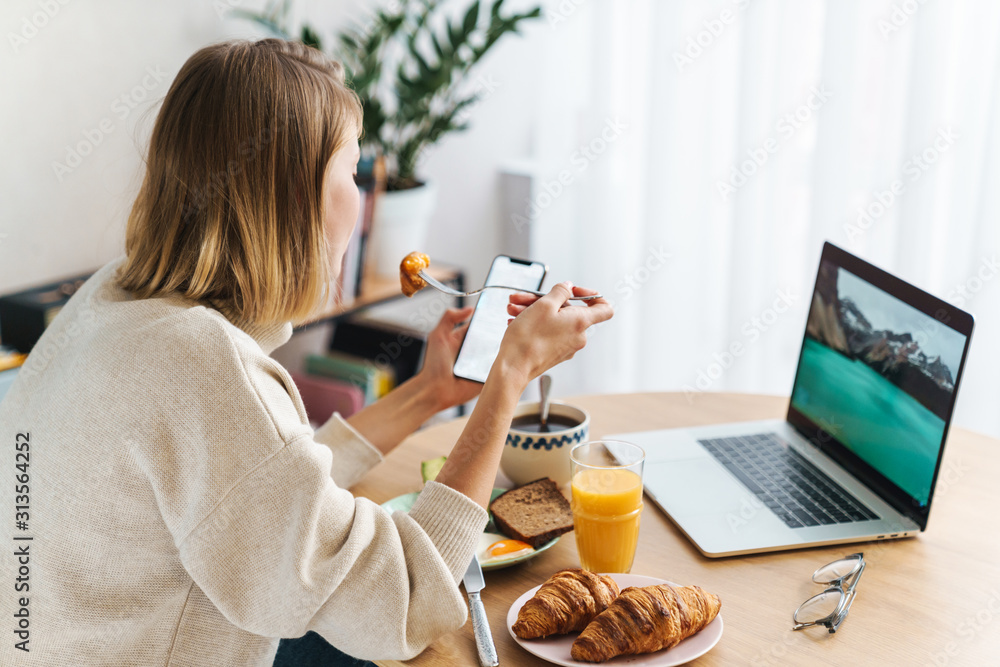 Photo of young woman using laptop and cellphone while having breakfast - obrazy, fototapety, plakaty 