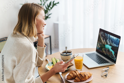 Photo of smiling woman using laptop and cellphone while having breakfast