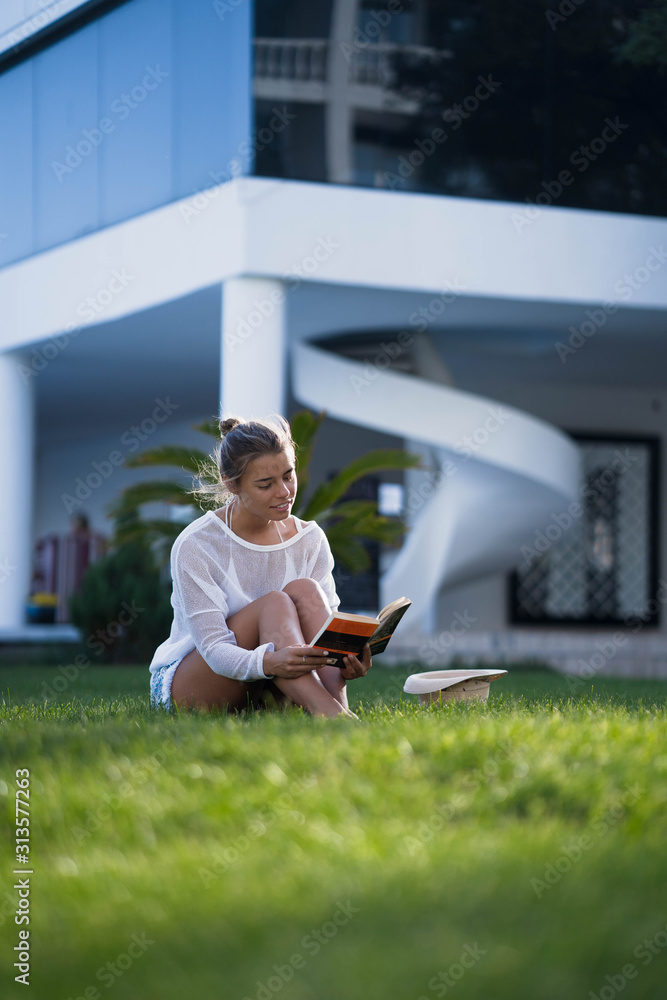 Beautiful smart girl with gathered hair sitting on a green lawn and reading book. Girl wearing in bikini and white tunic. The suns rays shining on her. Beautiful smart girl with gathered hair sitting on a green lawn and reading book. Girl wearing in bikini and white tunic. The suns rays shining on her.