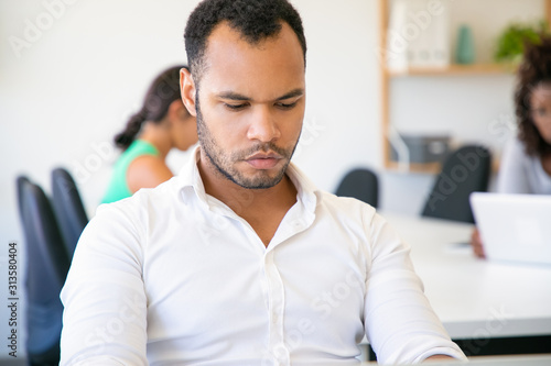 Pensive office worker sitting at table and looking at laptop. Front view of concentrated African American man working in office. Employment concept