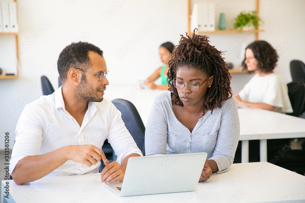 Two colleagues communicating during working process. Focused employees looking at each other while sitting at table with laptop. Teamwork concept Two colleagues communicating during working process. Focused employees looking at each other while sitting at table with laptop. Teamwork concept
