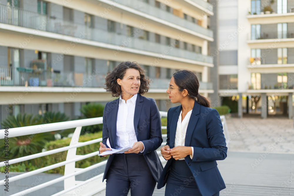 Serious female colleagues with documents discussing project on their way to office. Business women walking outside in city. Corporate communication concept Serious female colleagues with documents discussing project on their way to office. Business women walking outside in city. Corporate communication concept