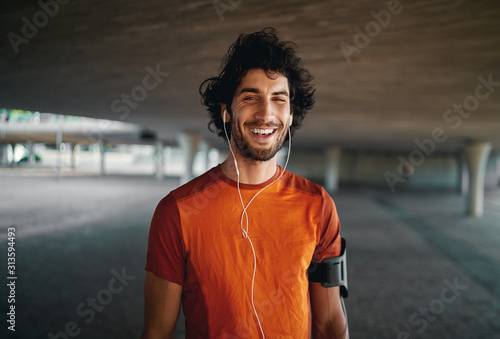 Cheerful portrait of a healthy sporty young man enjoying listening to music on earphones standing on city street