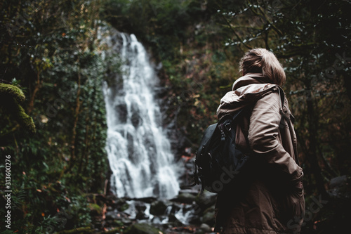girl with a backpack on her back on a path overgrown with trees and a moss in a dark gloomy forest in the rain and looks at a high powerful waterfall