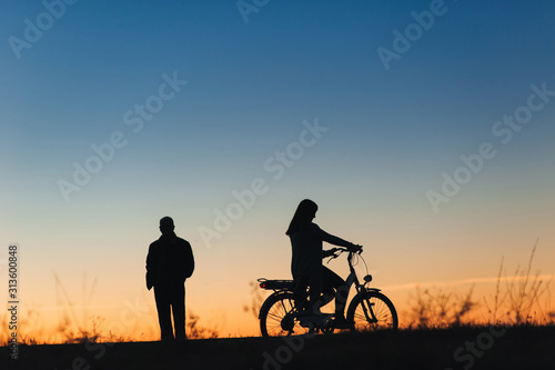 Female cyclist on the e-bike or electric bicycle on the sunset background. Silhouette of the woman and man in profile. Active leisure. Travel. Sport.