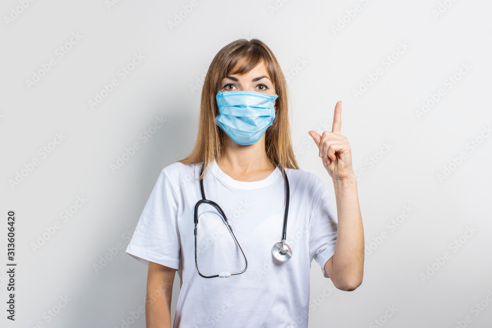 Young woman in medical mask and stethoscope pointing finger on a light background. Banner. Concept doctor, high level medical medicine, vaccination, visit to the doctor Young woman in medical mask and stethoscope pointing finger on a light background. Banner. Concept doctor, high level medical medicine, vaccination, visit to the doctor
