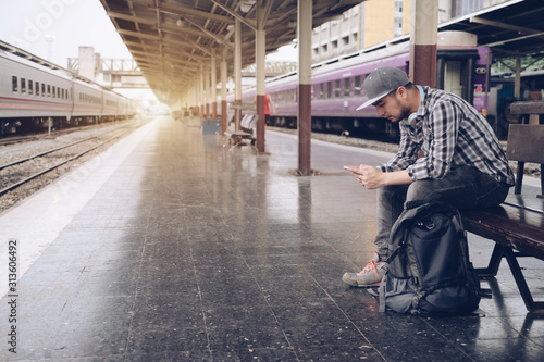 Young man using mobile smart phone checking train timetable at train station platform. Asian traveler text message with cellphone while waiting for train. Technology travel lifestyle concept.