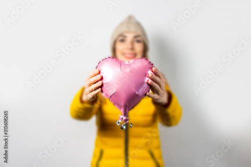 Young woman in a yellow down jacket and hat holds an air balloon in the shape of a heart on a light background. Valentine's day concept, gift, holiday, lover. Banner © Alex