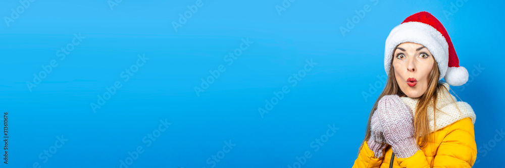 Young woman with a surprised face in a yellow jacket and a hat of Santa Claus on a blue background. Concept of the winter holidays, Christmas, New Year, surprise, shock. Banner Young woman with a surprised face in a yellow jacket and a hat of Santa Claus on a blue background. Concept of the winter holidays, Christmas, New Year, surprise, shock. Banner