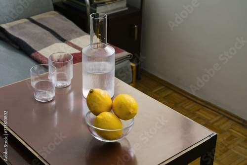 Three fresh yellow lemons in a glass bowl, two glasses and carafe with water on table, still life