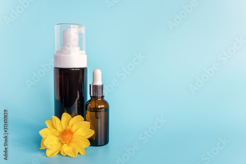 Essential oil in small open brown dropper bottle with lying glass pipette, big bottle with white dispenser and yellow flower on blue background. Concept natural organic beauty cosmetics product