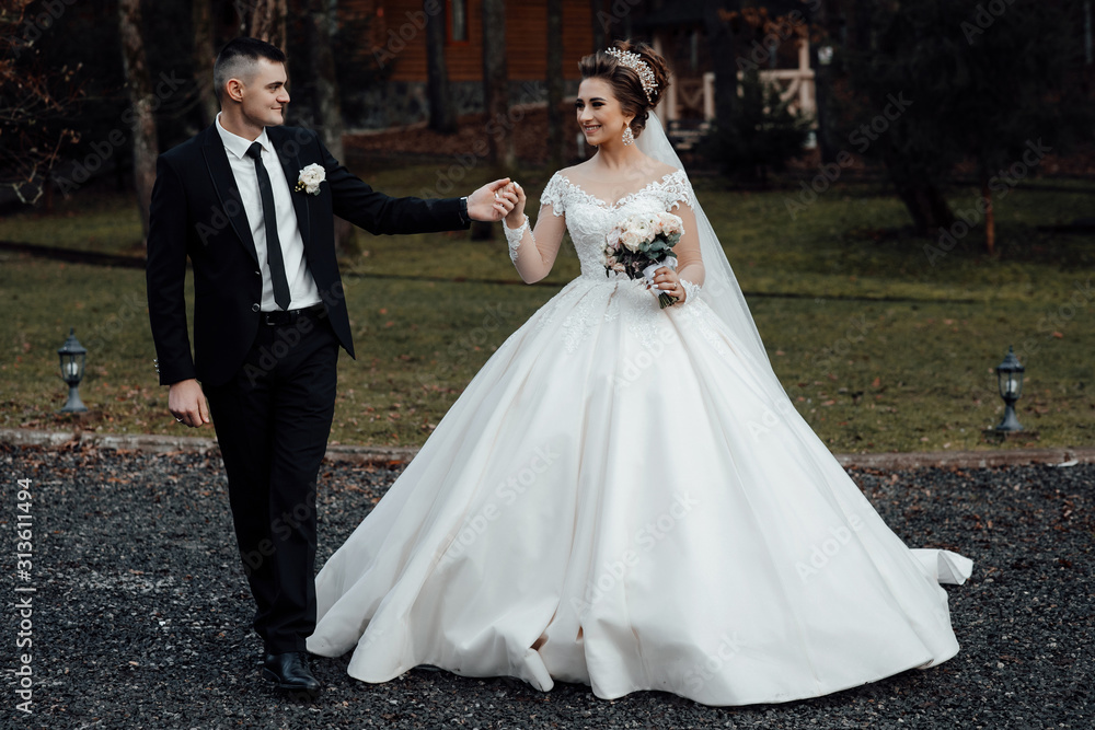 A young and beautiful bride and her husband is standing in park with bouquet of flowers. Amazing smiling wedding couple Beautiful bride and groom on their wedding day. Loving wedding couple outdoor. 