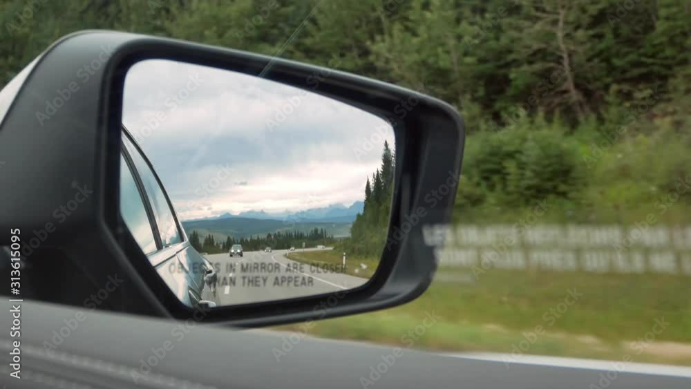 POV of side mirror of the moving car while while driving along the local natural way in canada with natural view of Mountain range and pine tree forest in summer holiday POV of side mirror of the moving car while while driving along the local natural way in canada with natural view of Mountain range and pine tree forest in summer holiday